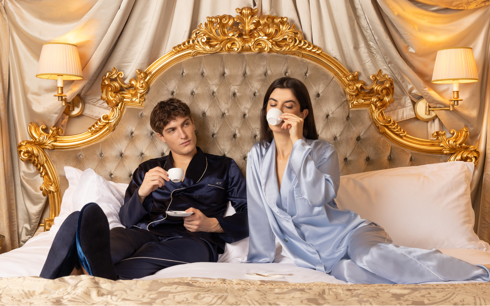 Couple in formal pajamas sitting on a luxurious bed with gold headboard.